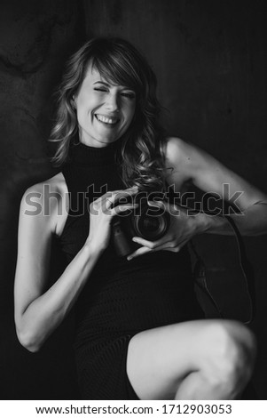 Beautiful young woman in a black dress with a camera in her hands in a loft studio winks and smiles. Soft selective focus. Woman photographer. Black and white art photo.