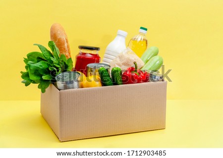 Donation Box with Supplies Food for People in Isolation on Yellow Background. Essential Goods: Oil, Canned Food, Cereals, Milk, Vegetables, Fruit Royalty-Free Stock Photo #1712903485