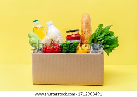 Donation Box with Supplies Food for People in Isolation on Yellow Background. Essential Goods: Oil, Canned Food, Cereals, Milk, Vegetables, Fruit Royalty-Free Stock Photo #1712903491