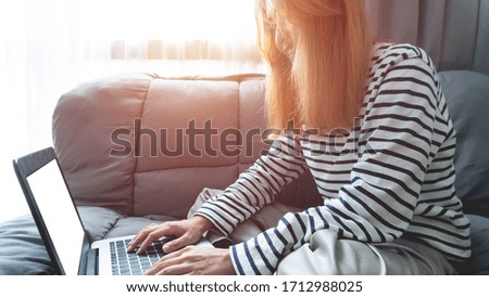 Smiling young women working from home or learning online education   on laptop computer while sitting on the grey sofa at the living room, while typing an email in during the Coronavirus or Covid-19.