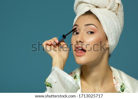 Young attractive funny woman putting eye mascara, studio shot over blue background, beauty treatment morning 