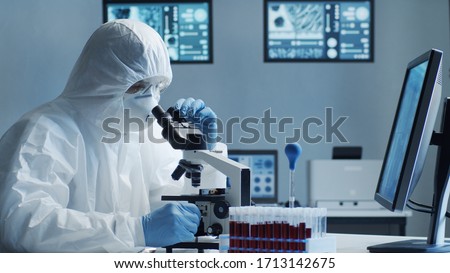 Scientist in protection suit and masks working in research lab using laboratory equipment: microscopes, test tubes. Coronavirus 2019-ncov hazard, pharmaceutical discovery, bacteriology and virology. Royalty-Free Stock Photo #1713142675
