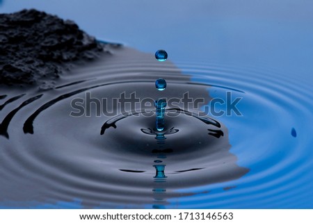 Blue colored water drops frozen in the air and forming ripples on the liquid surface, with rocks or stones in the background.