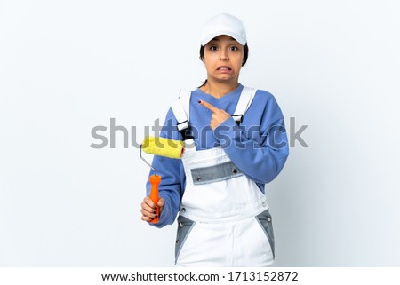 Painter woman over isolated white background frightened and pointing to the side