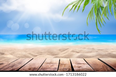 A wooden table product display with a summer vacation, holiday background of a tropical beach, blue sea, white clouds, sun flare and green palm tree leaves.