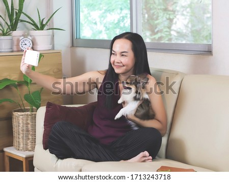 Beautiful Asian woman sitting on couch in living room taking selfie photo with her Chihuahua dog on lap , work from home , social distancing concept.