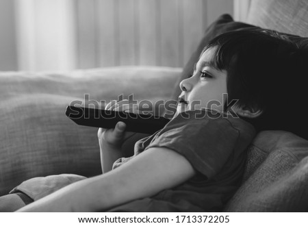 Black and White Kid lying on sofa holding remote control and watching cartoon on TV with morning light,Child boy stay at home relaxing in living room during covid 19 lockdown, Social Distancing