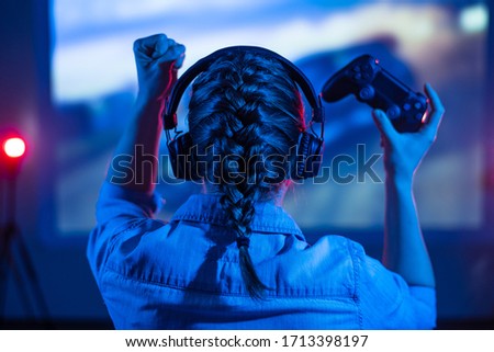 Gamer plays a video game on a TV or PC screen. Girl with a joystick and headphones in a neon room. Online gaming, simulation games. Tournaments between players. Dark background, back view.