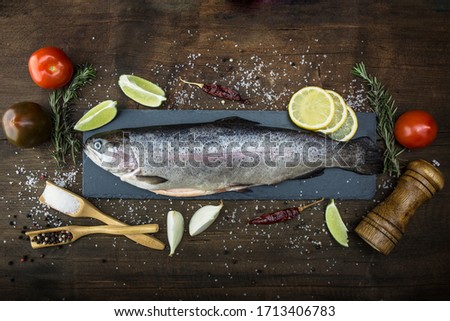 Raw trout lie on a black stone chopping Board on a wooden rustic table. Nearby are: lime, tomato, rosemary, lemon, pepper, onion, salt. Copy space. Royalty-Free Stock Photo #1713406783