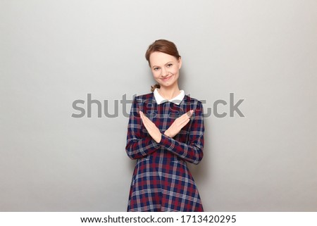 Studio portrait of happy cheerful blond girl wearing checkered dress, making cross with hands, showing stop, prohibition or enough gesture, smiling joyfully, standing over gray background
