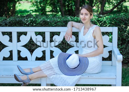 Young beautiful woman in a white wedding dress is sitting on a white bench in the summer park.