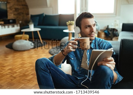 Smiling man using digital tablet and listening music over headphones while drinking coffee at home.