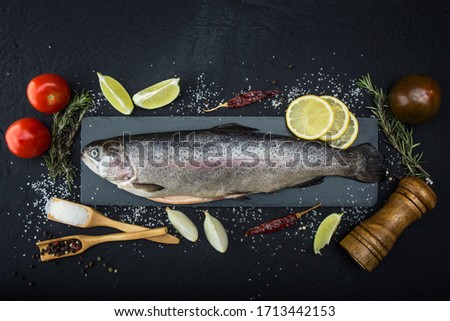 Raw trout lie on a black stone chopping Board on a dark concrete table. Nearby are: lime, tomato, rosemary, lemon, pepper, onion, salt. Copy space. Royalty-Free Stock Photo #1713442153