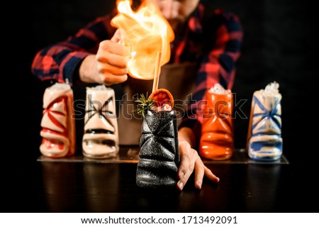 Close-up professional bartender accurate decorates colored ceramic glasses with ice by slice of citrus and make fire over them