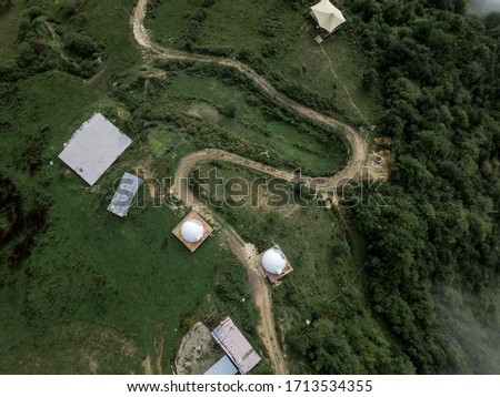 Top down aerial drone view from above of curved countryside road in the forest in mountains and small agricultural farmer settlement with buildings and yurts. Rural Tibetan area of Sichuan province.