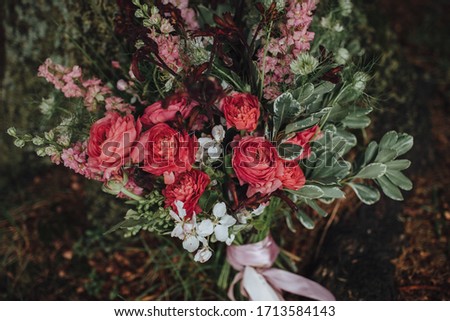 Wedding floristry. A bouquet of red and white flowers with greenery and a pink ribbon stands on the ground near a tree covered with moss.