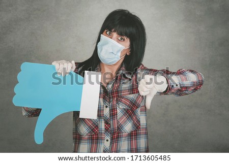 young woman wearing medical mask for coronavirus holding a dislike icon over gray background