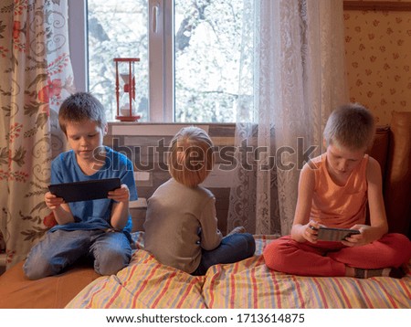 Three boys are playing with tablet and smartphone on leather sofa in bedroom by window. brothers spend time together in quarantine in rural house. Older children take pictures of young and show photos