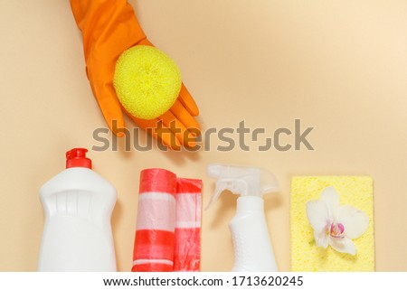 Woman's hand in an orange protective glove holding a sponge with a washing and cleaning set on the beige background. Top view.