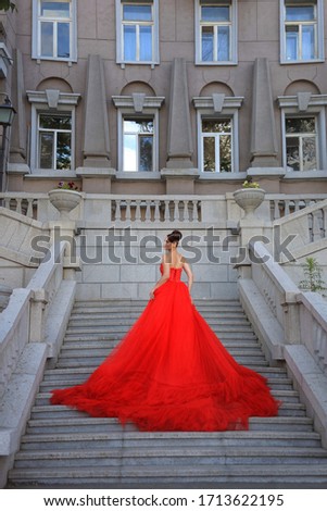 beautiful girl with a beautiful hairstyle in a long red dress stands on the stairs of a gray historic building
