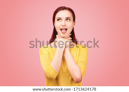 Funny girl makes faces hands behind neck, tongue sticking out, eyes bulging. The concept of frivolous fun and teasing. Young girl dressed in yellow sweater stands isolated on pink background in Studio
