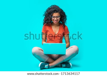 Beautiful young smiling woman in casual outfit and trendy eyeglasses sitting isolated on bright colored blue background and working on her laptop. Full length horizontal studio shot.  Royalty-Free Stock Photo #1713634117
