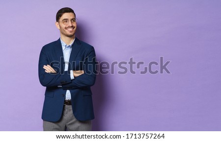 Horizontal shot of young business male standing in jacket and eyeglasses with arms crossed, looking aside with positive smile, isolated on purple background, copyspace on right