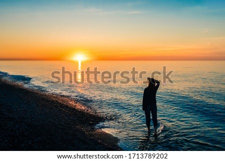A silhouette lonely person ankle-deep in the sea and looks the sunset. Trousers rolled up to the knee. Bright orange colors of the sunset sky on the horizon. Calm sea. Light waves roll in on the fores