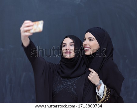 group of young beautiful muslim women in fashionable dress with hijab using mobile phone while taking selfie picture in front of black chalkboard representing modern islam fashion technology 