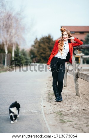 red-haired jockey girl in a red cardigan and black high boots with a cat for a walk