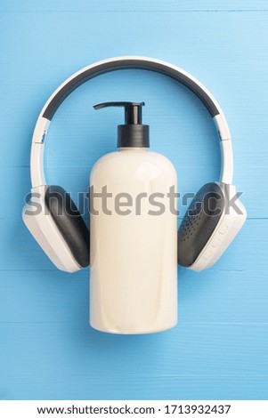 Plastic beige gel soap container with dispenser and white black wireless headphones on a light blue wooden background. The concept of coronavirus stay at home