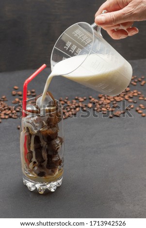 Hand pours milk from measuring cup into glass cup with coffee ice cubes. Grains of coffee on black background. Close up.
