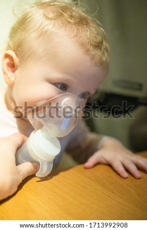 Cute little baby in mask of an inhaler. Procedure of inhalation at home. Baby taking respiratory therapy with nebulizer. Watching cartoons on your phone.