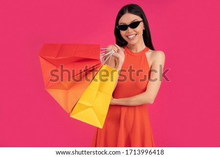 Image of a beautiful excited happy asian woman posing isolated over pink wall background holding shopping bags. dressed in orange dress and sunglasses.