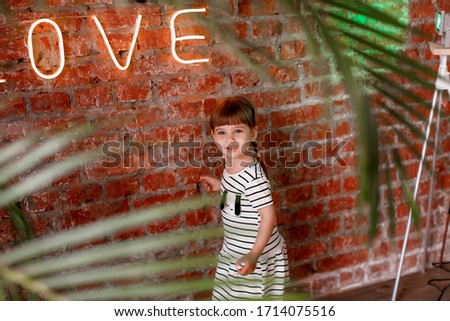 Neon inscription on the wall, shot through palm leaves, decor element. A girl stands near the wall, smiling