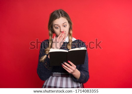 schoolgirl teen with two pigtails in a jumper and checkered skirt with a book in hand on a red background.