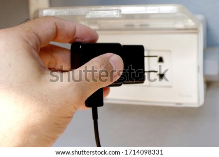 Close up left hand of man is holding black plug and prepare for battery charging a mobile phone on the electric socket on the gray color wall, selective focus picture