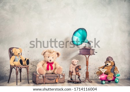 Antique gramophone phonograph, and retro Teddy Bear toys on chair, old luggage, wooden horse, plane front concrete wall background. Listening to nostalgic music concept. Vintage style filtered photo