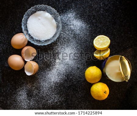 Ingredients for a lemon Meringue Tart, including eggs, sugar, lemon, and a can on condense milk, on a dark background