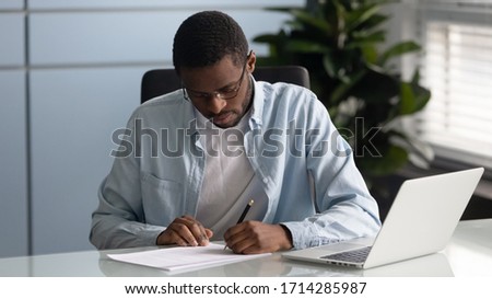 Focused african american businessman writing signing paper document. Serious diverse man puts signature on business document, agreement, using laptop in office, serious employee making notes on paper.