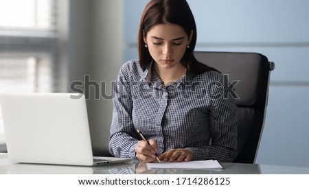 Young serious businesswoman sign up document at work. Tired woman employee writing report on paper near modern laptop. Success business deal concept. Royalty-Free Stock Photo #1714286125