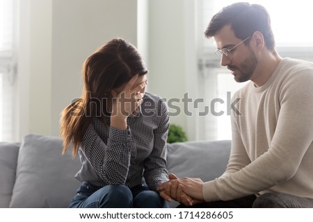 Handsome husband comforting wife, holding hands, showing support. Man upset wife, expressing sympathy and understanding, say sorry, ask for forgiveness, family sitting on comfortable sofa at home.