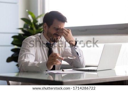 Stressful businessman taking off glasses, suffering from dry eyes syndrome after long laptop use. Exhausted employee executive manager ceo feeling eye strain, massaging nose bridge Royalty-Free Stock Photo #1714286722