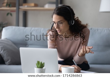 Smart young woman in glasses wearing wireless headphones with mic, looking at laptop monitor. Focused millennial female student watching online educational webinar, writing notes, studying at home. Royalty-Free Stock Photo #1714286863