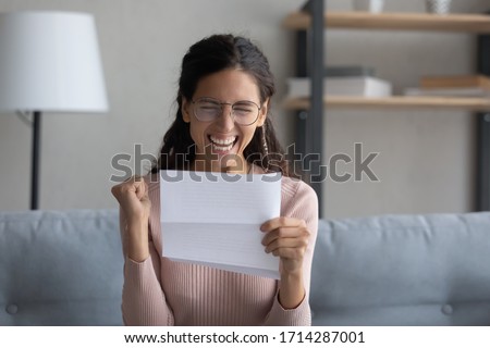 Overjoyed young attractive lady in eyewear holding paper letter, celebrating business success. Emotional millennial woman making yes gesture, feeling excited about personal achievement at home. Royalty-Free Stock Photo #1714287001