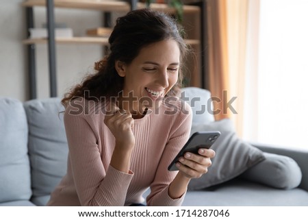 Joyful millennial woman sitting on couch, reading sms with good luck news. Happy young lady celebrating online lottery win, enjoying communicating with friends or beloved man, feeling excited at home. Royalty-Free Stock Photo #1714287046