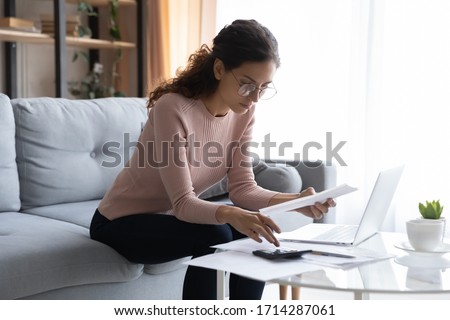 Focused young woman in glasses holding paper documents invoices, calculating business or household expenses, managing monthly budget incomes or outcomes, planning investments alone in living room. Royalty-Free Stock Photo #1714287061