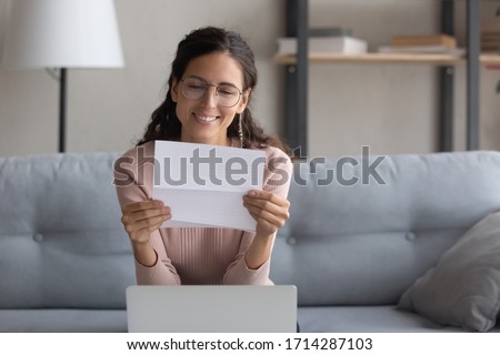 Smiling pretty millennial female student in glasses sitting on sofa, reading paper university admission notification. Happy pleasant young woman wearing eyeglasses reading good news letter at home. Royalty-Free Stock Photo #1714287103