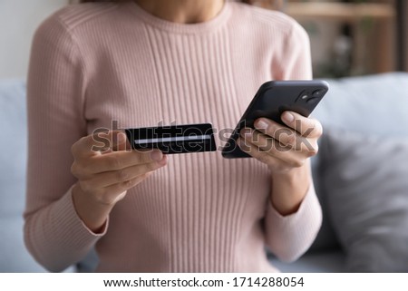 Close up young woman holding plastic credit card in one hand and telephone in another, entering cvv code, confirming purchase in online store. Female client using e-banking application on smartphone. Royalty-Free Stock Photo #1714288054