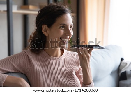 Head shot attractive smiling millennial woman recording voice message on smartphone. Happy young lady activating mobile virtual assistant, communicating with friends online, relaxing on cozy couch.